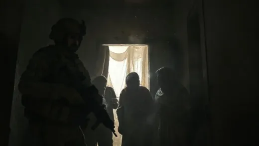 A tense scene inside a dimly lit house in Anbar, Iraq. US soldiers in combat gear are carefully securing local residents, who appear startled and frightened. One soldier holds a weapon, another interacts with a local, trying to calm them. Dust motes visible in the air, creating a dramatic atmosphere.
