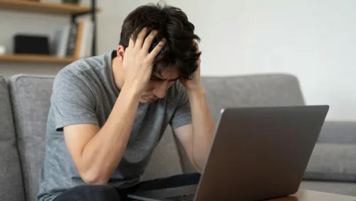 A person sitting on a couch at home, looking stressed while checking a smartphone and laptop. The room has a blurred background, suggesting work-life blending.