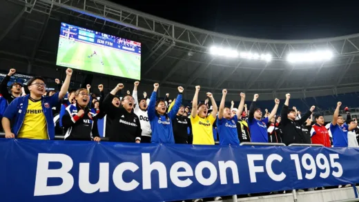 A vibrant and dynamic K-League soccer stadium scene at night, filled with passionate fans cheering for Bucheon FC 1995. The stadium lights are bright, showcasing a scoreboard celebrating Bucheon's promotion. In the foreground, a prominent banner with "Bucheon FC 1995" is visible. There are subtle hints of rivalry with another team (Anyang FC) in the background, perhaps a blurred glimpse of a rival team's color. The overall atmosphere is energetic and triumphant.