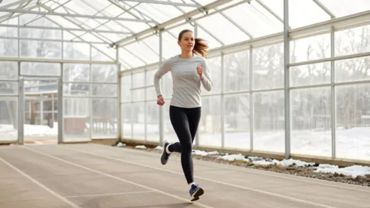 An energetic female runner, wearing a light long-sleeved shirt and leggings, running with a determined expression inside a large, transparent greenhouse track. The greenhouse interior is brightly lit by natural light, and outside, subtle hints of a cold winter landscape can be seen. The track is clearly marked. Focus on the runner's movement and the warm, inviting atmosphere contrasted with the cold exterior.