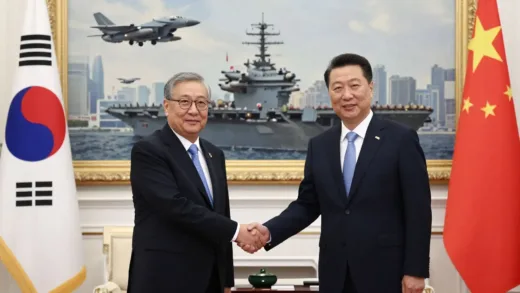 An image showing a formal handshake between Korean and Chinese leaders in a grand hall, with a superimposed, slightly transparent image of US fighter jets flying over a city or a US aircraft carrier in the background, symbolizing a hidden threat or concurrent event.