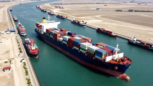 An impactful aerial shot of the Ever Given container ship stuck sideways in the Suez Canal, with many other ships waiting in queue. The scene should convey a sense of global paralysis and vulnerability of supply chains.