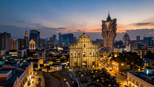 A panoramic view of Macau, showing both historic European-style buildings and modern skyscrapers with dazzling night lights. The sky is a blend of sunset colors.