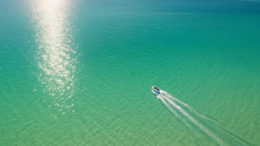 A wide aerial view of the shallow, turquoise Sea of Azov with a small boat leaving a trail, sunlight reflecting on the calm water surface.