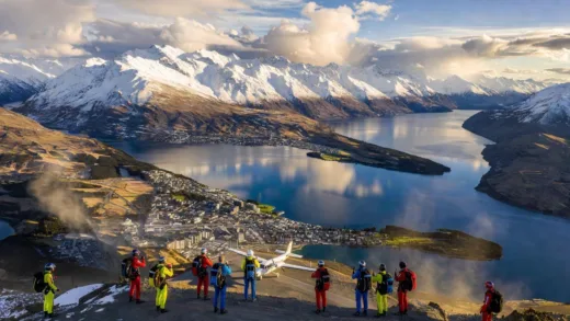 A breathtaking aerial view of Queenstown, New Zealand with snow-capped mountains, Lake Wakatipu, and adventure seekers preparing for skydiving. The sky is partly cloudy with dramatic lighting.