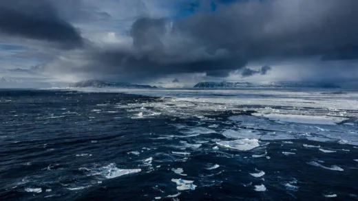 An aerial view of the vast and icy Bering Sea, showing the harsh conditions and the distant, rugged coastlines of Alaska and Chukotka. The water is a dark, cold blue, with hints of ice formations. Dramatic clouds are in the sky.