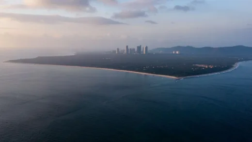 A dramatic aerial shot of a vast, empty coastline in Angola, with a distant hint of modern high-rise buildings of Luanda on the horizon, under a slightly misty sky at dawn.
