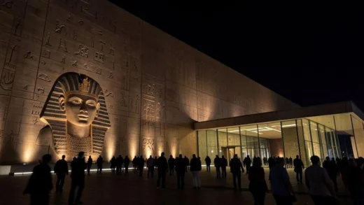 Grand Egyptian Museum exterior at night with dramatic lighting, showing modern architecture alongside ancient Egyptian motifs, with crowds of visitors entering