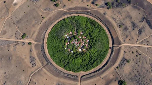 A satellite image of a perfectly circular geological formation in Madagascar, with a small, densely populated village clearly visible in its lush green center, surrounded by contrasting barren landscapes.