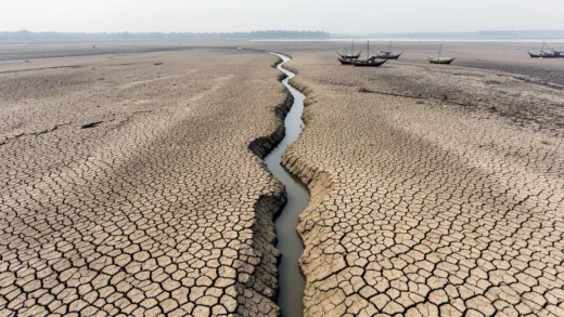 An aerial view of a wide, once-mighty riverbed in Southeast Asia, now largely dry and cracked earth, with a small, winding stream in the center. In the distance, traditional fishing boats are stranded. The sky is hazy. Focus on the scale of dryness.