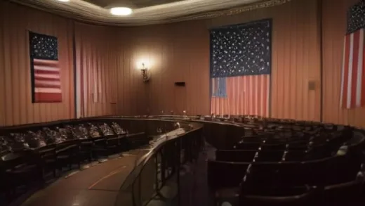 A dramatic wide-angle illustration of a U.S. congressional hearing room, an elderly scientist in a suit seated at a long table with a microphone, rows of microphones and American flags in the background, warm documentary photography style, tense atmosphere, soft depth of field