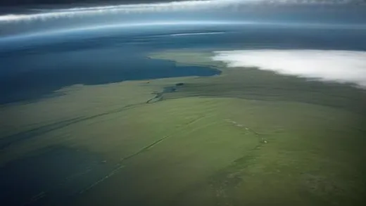 Aerial view of the windswept Falkland Islands coastline with vast green plains dotted with thousands of white sheep, dramatic storm clouds, a distant military radar station and runway, cinematic wide shot, moody South Atlantic atmosphere